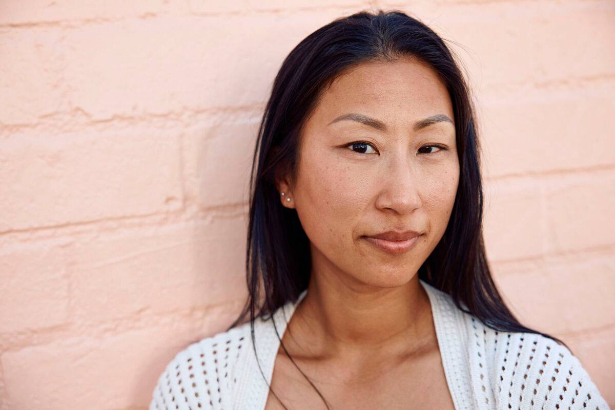 Close-up portrait of a person with long dark hair standing against a light pink wall, wearing a light-colored top.